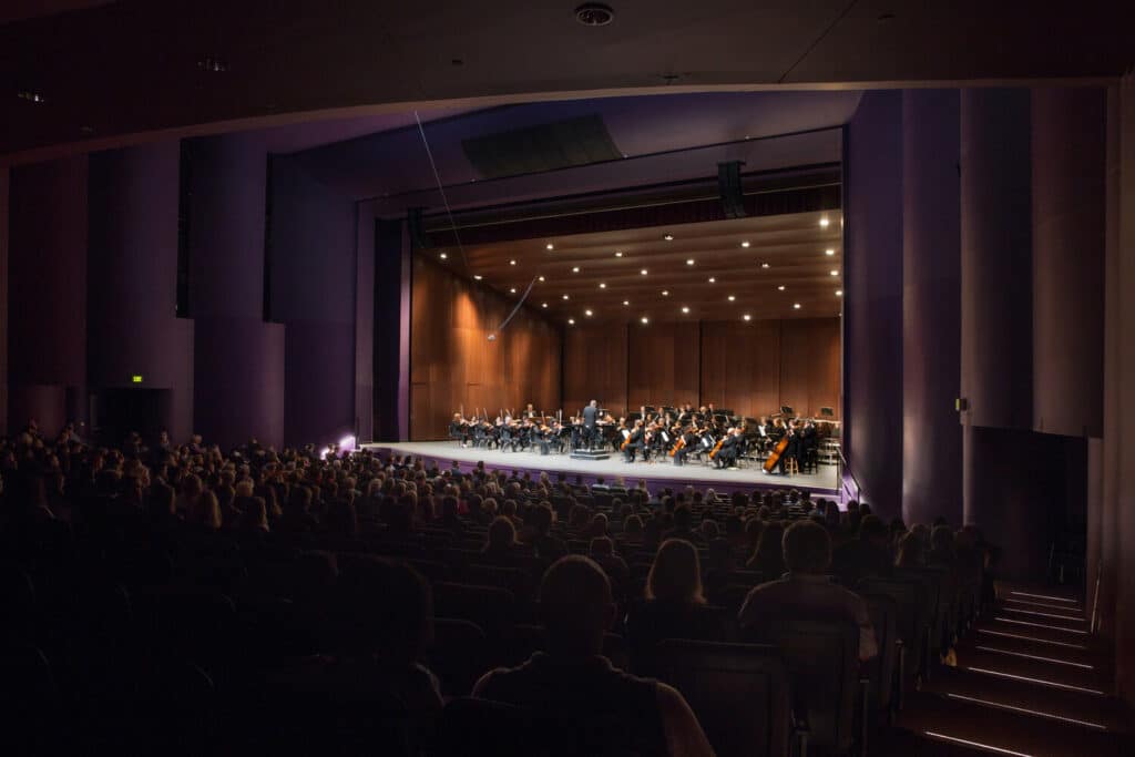 An orchestra concert in an auditorium. The audience is full of attendees.